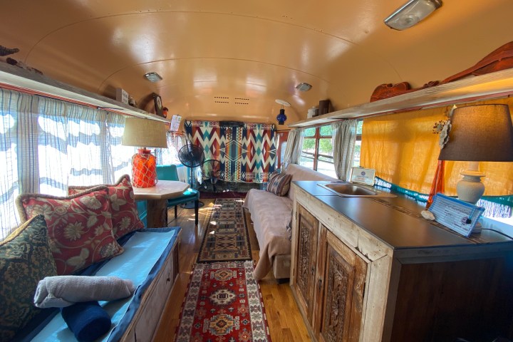 Cozy bus interior with colorful cushions, decorative rugs, and wooden furnishings, featuring natural light from windows.