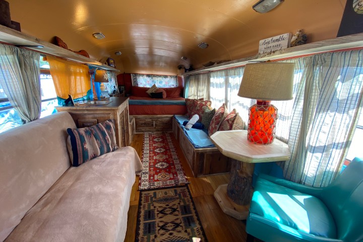 Cozy bus interior with couches, bed, colorful pillows, and a red lamp on a wooden table.