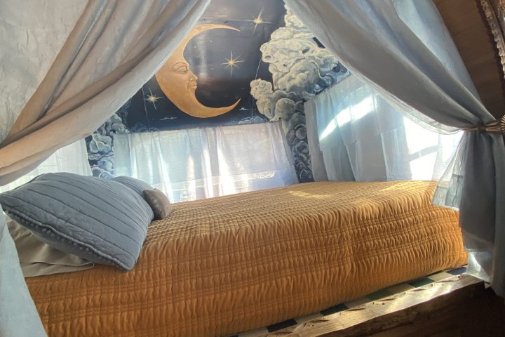 Cozy bed with orange quilt, blue pillows, curtains, and moon mural on wall.