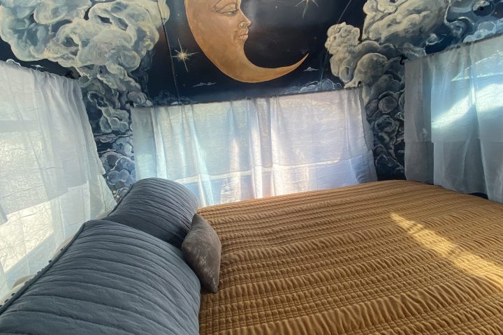 Bed with a blue and orange cover beneath a ceiling mural of moon, stars, and clouds.
