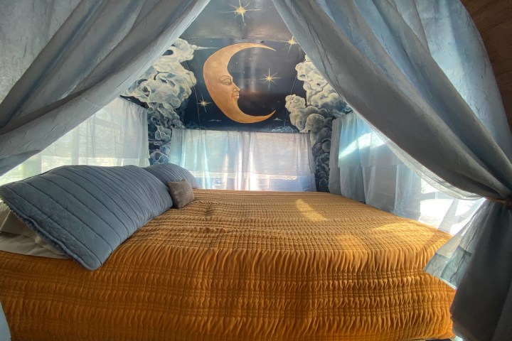 Cozy bed with canopy, blue pillows, and moon-themed mural on wall.